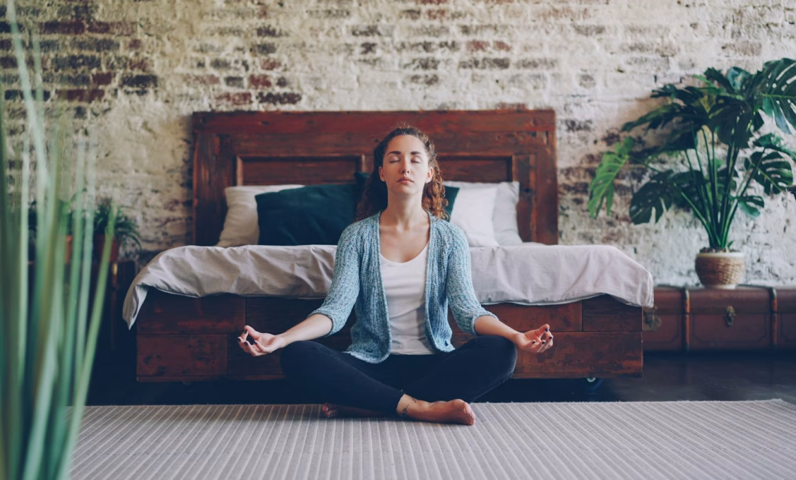 Woman sitting cross-legged in a calm, sunlit bedroom, practising relaxation at home for IBS bloating