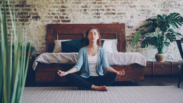 Woman sitting cross-legged in a calm, sunlit bedroom, practising relaxation at home for IBS bloating